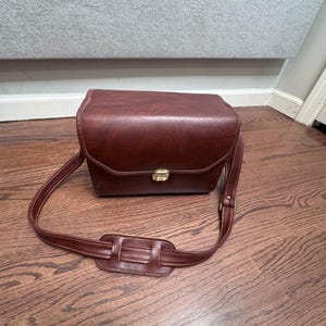 Puede incluir: Bolso de cámara vintage rectangular en un rico color marrón rojizo. El bolso presenta una solapa superior con un cierre dorado y una correa para el hombro a juego con una sección acolchada para mayor comodidad. El bolso está hecho de un material similar al cuero.