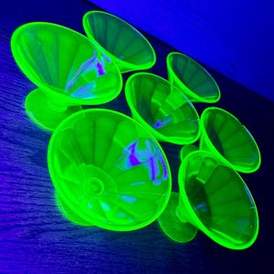 Può includere: Una collezione di coppette da dessert in vetro verde brillante, in stile vintage. Le coppette hanno una base a piedistallo e un design a coppa scanalata. Il vetro brilla con una tonalità verde neon su uno sfondo blu scuro. La disposizione suggerisce un set di pezzi da portata.