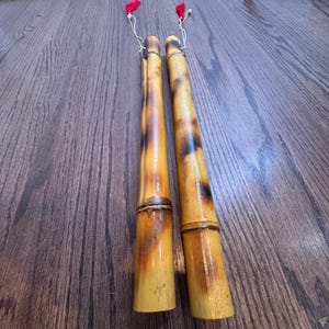 Peut inclure: Deux claquettes en bambou avec une finition naturelle et des accents brun foncé. Chaque claquette a un pompon rouge attaché au sommet avec une ficelle blanche. Les claquettes sont posées sur une surface en bois.
