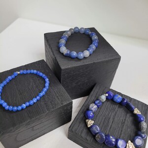 May include: Three bracelets made of blue gemstone beads displayed on black wooden stands. The bracelets are different lengths and feature different styles of beads.