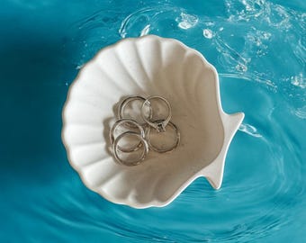 Puede incluir: Un plato de cerámica blanca en forma de concha de mar con cuatro anillos plateados en su interior. El plato flota en una piscina de agua azul.