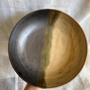 Puede incluir: Un cuenco de cerámica redondo con un esmalte de dos tonos. Una mitad del interior es de color negro carbón liso, mientras que la otra mitad es de color beige terroso moteado. Una banda vertical de verde oliva apagado separa las secciones.