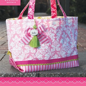 Może przedstawiać: Różowa i biała torba typu tote z wzorem damaskowym. Torba ma zielony frędzl i różową i białą wstążkę w paski z etykietą z napisem "bébé". Torba leży na chodniku z cegły. Tekst "the out n about tote" i "A multi-purpose tote for today's life on the go!" znajduje się na dole obrazu.
