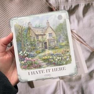 May include: A clear tablet case featuring a watercolor painting of a cottage with a stone path and colorful flowers. The text "I HATE IT HERE" is printed at the bottom, with "TTPD" below it.