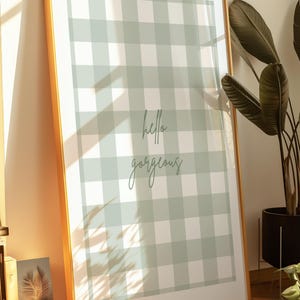 May include: A framed print with a gold border. The artwork features a light green and white gingham pattern with the words "hello gorgeous" in a script font. The print is leaning against a wall.