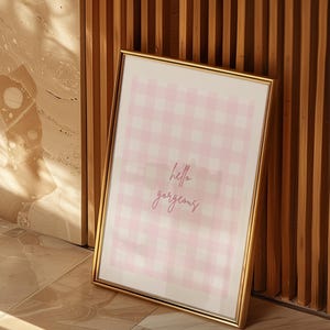 Op de afbeelding: Een ingelijste print met een gouden rand, leunend tegen een houten muur. De print heeft een roze en wit ruitpatroon met de woorden "hello gorgeous" in een cursief lettertype. De print wordt weergegeven op een betegelde vloer.