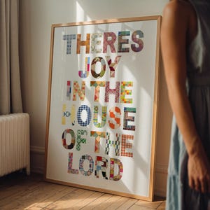 May include: A framed art print with the words "THERES JOY IN THE HOUSE OF THE LORD" in colorful patchwork letters. The artwork is in a light wooden frame and leans against a wall. The print is on a white background.