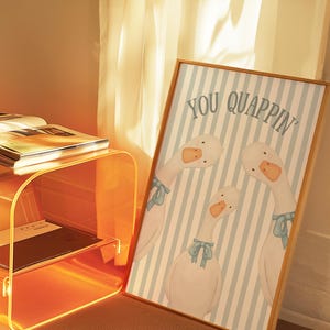 May include: A framed art print featuring three white geese with blue bows against a light blue and white striped background. The words "YOU QUAPPIN'" are printed at the top. The artwork is displayed in a room with an orange acrylic side table.
