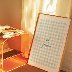 May include: A framed print with a light blue and white gingham pattern and the words "hello gorgeous" in cursive. The print is leaning against a wall next to a clear orange acrylic side table.