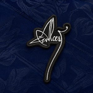 May include: A dark blue fabric featuring a tonal floral jacquard pattern with a black embroidered patch centred on the surface. The patch displays a white stylised signature or logo that reads Alces in a cursive script with a long trailing line.