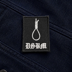 Op de afbeelding: Een rechthoekige zwarte geborduurde patch met een wit gestikt strop-icoon boven de tekst DSBM in een gotisch lettertype. De patch is bevestigd op een donker marineblauwe stoffen ondergrond met een zichtbare horizontale ribbelstructuur en een metalen knoop.