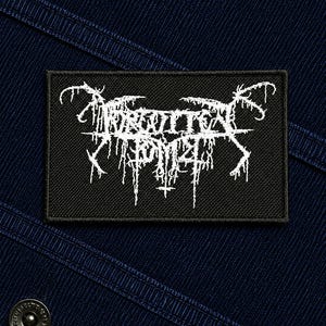 May include: A rectangular black embroidered patch featuring white text in a stylized, jagged, and dripping font. The patch is attached to a dark blue fabric surface that includes a visible metal button with circular text detailing.
