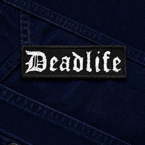 Peut inclure: Un écusson rectangulaire noir brodé avec le mot Deadlife en lettres blanches de style gothique. L'écusson est cousu sur un tissu en jean bleu foncé avec des coutures visibles et un bouton métallique visible dans le coin inférieur gauche.