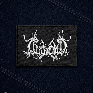 May include: A rectangular black fabric patch featuring white embroidered text in a stylized, intricate, and jagged font. The patch is centered on a dark navy blue denim fabric background with visible diagonal stitching and a single metal button.