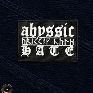 Puede incluir: Un parche rectangular negro bordado con texto en blanco. La línea superior dice "abyssic" en una fuente gótica, la línea central presenta caracteres de estilo rúnico y la línea inferior dice "HATE" en una fuente gótica en negrita.