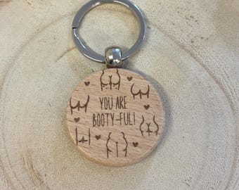 Könnte beinhalten: Ein Holzschlüsselanhänger mit einem silbernen Ring. Der runde Holzanhänger ist mit mehreren Gesäßillustrationen und dem Satz "YOU ARE BOOTY-FUL!" graviert. Der Schlüsselanhänger ist aus hellem Holz gefertigt.