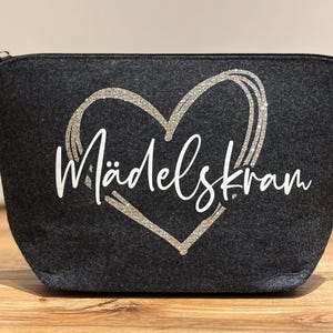 Könnte beinhalten: Dunkelgraue Kosmetiktasche mit Reißverschluss, verziert mit einem glitzernden Herz und dem Schriftzug "Mädelskram" in Weiß. Die Tasche besteht aus einem weichen Material und ist für Kosmetika geeignet.