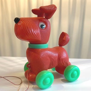 Può includere: Un giocattolo vintage da tirare, un cane in plastica rossa con ruote verdi e un collare verde. Il cane ha un naso nero, occhi mobili e un cordino. Un classico giocattolo per bambini.