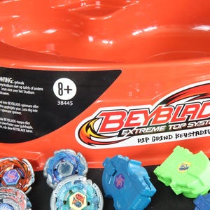 May include: An orange Beyblade Extreme Top System stadium with several colorful Beyblade tops and launchers. The stadium has the text "Beyblade" and "Extreme Top System" printed on it. The image also includes the text "8+".