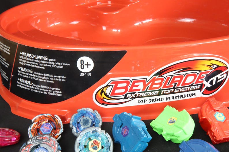May include: An orange Beyblade Extreme Top System stadium with several colorful Beyblade tops and launchers. The stadium has the text "Beyblade" and "Extreme Top System" printed on it. The image also includes the text "8+".