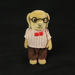 May include: A flocked dog figurine with tan fur, wearing round black glasses, a white and brown vertical striped short-sleeved shirt, a small red bow tie at the collar, and solid brown trousers. The figure has a smooth texture and upright posture.
