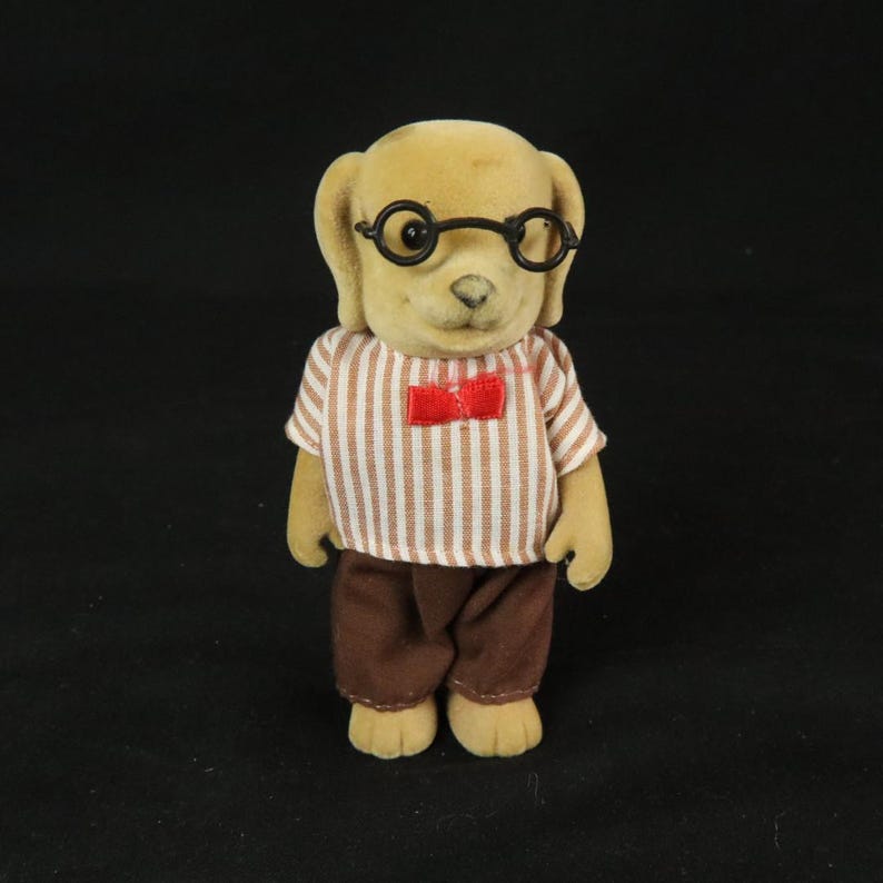 May include: A flocked dog figurine with tan fur, wearing round black glasses, a white and brown vertical striped short-sleeved shirt, a small red bow tie at the collar, and solid brown trousers. The figure has a smooth texture and upright posture.