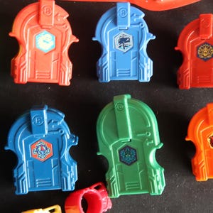 May include: A collection of colorful plastic toy components in red, blue, green, and orange. Each piece has a unique design with a central emblem. The toys appear to be part of a larger set, possibly for a game or construction activity.