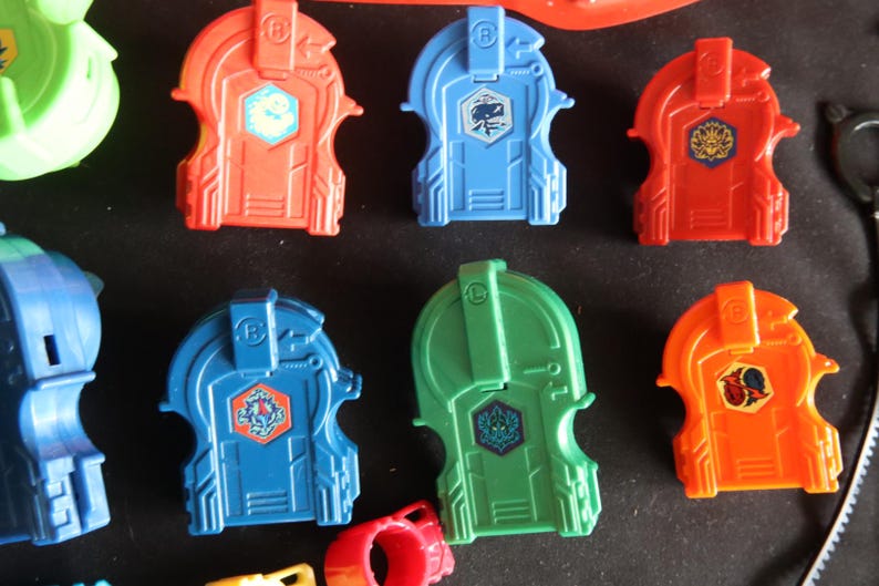 May include: A collection of colorful plastic toy components in red, blue, green, and orange. Each piece has a unique design with a central emblem. The toys appear to be part of a larger set, possibly for a game or construction activity.
