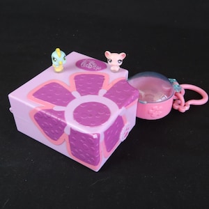 May include: A light purple toy box with a large flower design in shades of purple and pink. Two small animal figurines sit on top. A pink, round container with a clear top and a pink keychain is attached to the box.