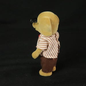 May include: A tan flocked animal figurine wearing a short-sleeved shirt with thin brown and white vertical stripes and solid brown trousers. The figure has black plastic glasses perched on its head and is viewed from a side profile angle.