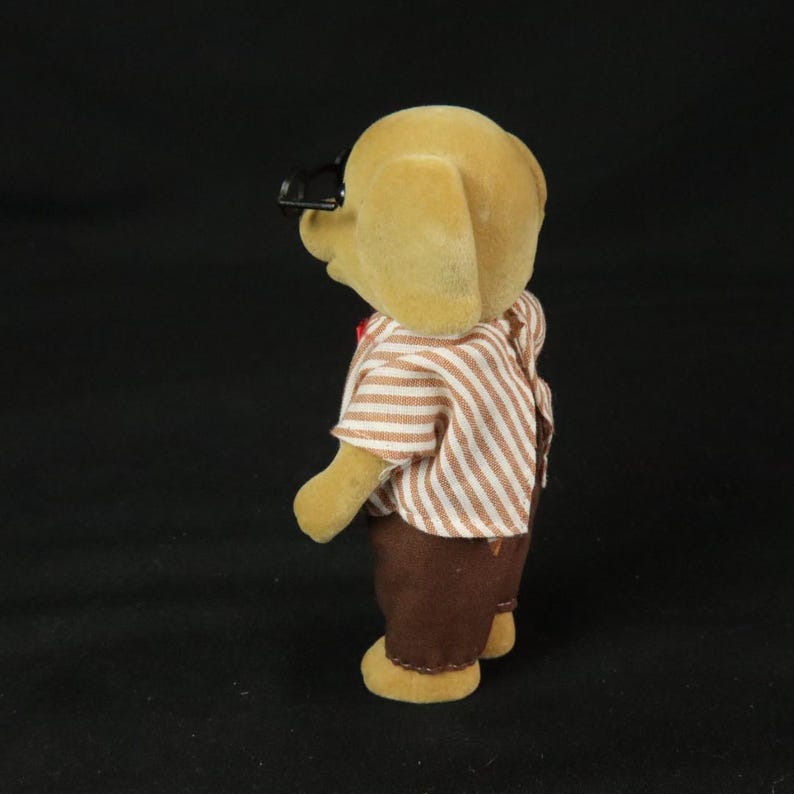 May include: A tan flocked animal figurine wearing a short-sleeved shirt with thin brown and white vertical stripes and solid brown trousers. The figure has black plastic glasses perched on its head and is viewed from a side profile angle.