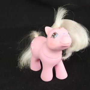 Op de afbeelding: Een roze plastic speelgoedpony met wit synthetisch haar op de manen en staart. De pony heeft grote blauwe ogen, een glimlachende uitdrukking en een symbool op de flank bestaande uit drie roze harten in een groepje. Het figuurtje staat rechtop.