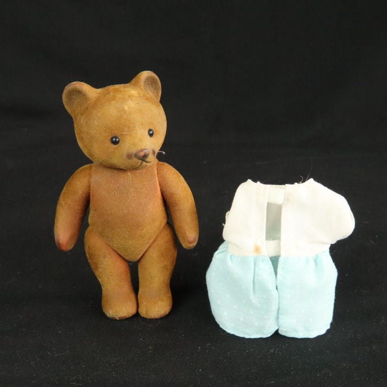 Peut inclure: Une petite figurine d'ours marron avec des yeux sombres et un nez cousu, plac&eacute;e &agrave; c&ocirc;t&eacute; d'un v&ecirc;tement s&eacute;par&eacute; compos&eacute; d'un haut blanc et d'une jupe bleu clair avec un motif discret &agrave; pois blancs, le tout sur un fond noir uni.