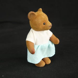 Peut inclure: Une petite figurine d'ours brun floqu&eacute; debout. L'ours porte un haut blanc &agrave; manches courtes associ&eacute; &agrave; une jupe bleu clair orn&eacute;e d'un motif subtil de petits points blancs r&eacute;p&eacute;t&eacute;s sur toute la surface du tissu.