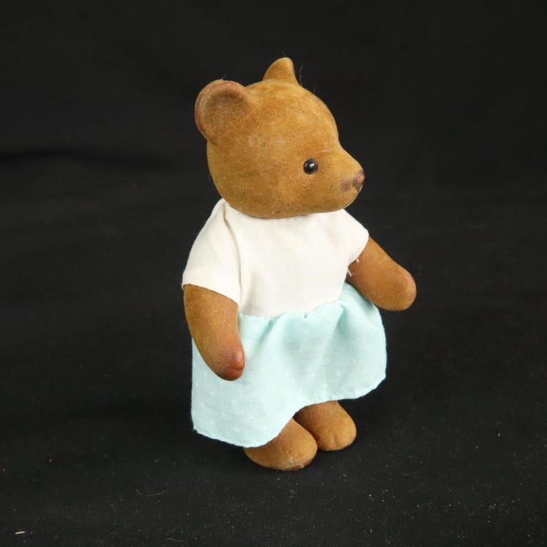 Peut inclure: Une petite figurine d'ours brun floqu&eacute; debout. L'ours porte un haut blanc &agrave; manches courtes associ&eacute; &agrave; une jupe bleu clair orn&eacute;e d'un motif subtil de petits points blancs r&eacute;p&eacute;t&eacute;s sur toute la surface du tissu.