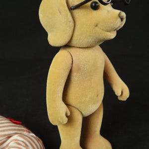 May include: A tan, flocked dog figurine standing upright with black, round-rimmed glasses perched on its head. The dog has small black eyes and a black nose. Beside the figurine lies a piece of white and brown striped fabric with a red ribbon.