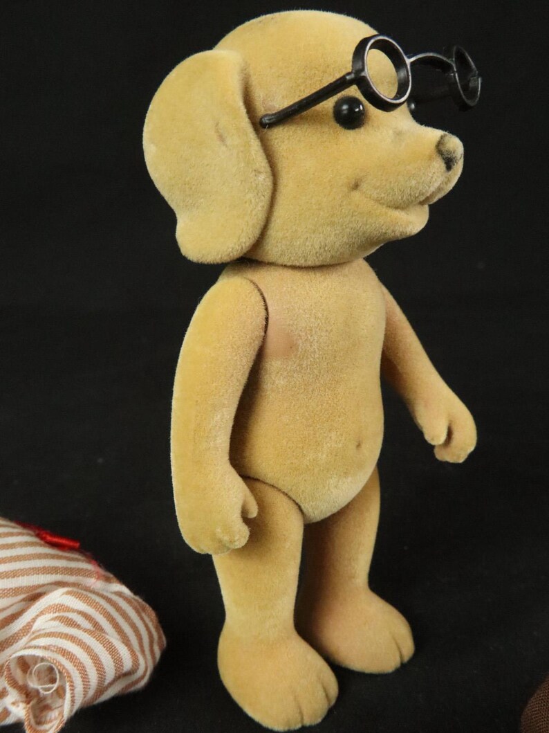 May include: A tan, flocked dog figurine standing upright with black, round-rimmed glasses perched on its head. The dog has small black eyes and a black nose. Beside the figurine lies a piece of white and brown striped fabric with a red ribbon.