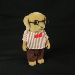 May include: A flocked dog figurine with tan fur, wearing round black-rimmed glasses, a short-sleeved shirt with vertical white and tan stripes, and a small red bow tie. The figure is dressed in solid brown trousers and has a neutral facial expression.