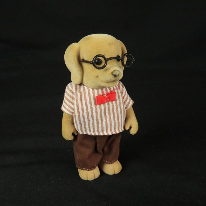 May include: A flocked dog figurine with tan fur, wearing round black-rimmed glasses, a short-sleeved shirt with vertical white and tan stripes, and a small red bow tie. The figure is dressed in solid brown trousers and has a neutral facial expression.