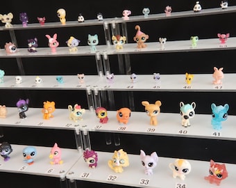May include: A multi-tiered clear acrylic display stand holding a collection of small, colorful plastic animal figurines. Each shelf features numbered labels in black ink, showcasing a variety of stylized creatures with diverse colors and poses.