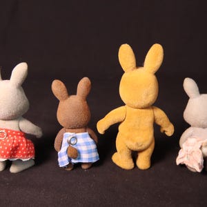 Może przedstawiać: Cztery figurki zabawek kr&oacute;lik&oacute;w w r&oacute;żnych kolorach. Jeden jest ż&oacute;łty, dwa szare, a jeden brązowy. Kr&oacute;liki mają na sobie sp&oacute;dnice lub ogrodniczki w czerwone grochy, niebiesko-białą kratę i r&oacute;żowe wzory kwiatowe. Zabawki stoją prosto.