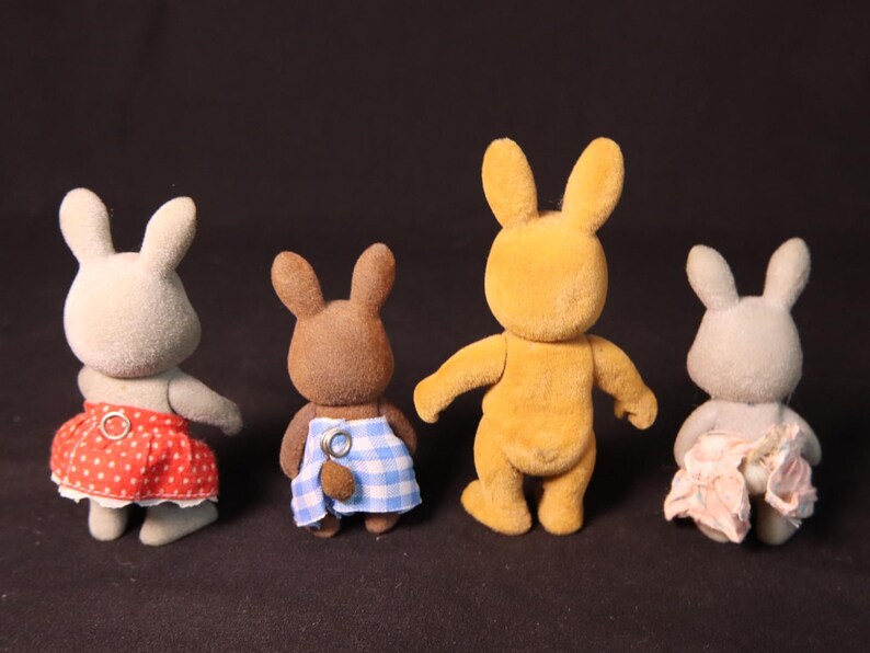 Może przedstawiać: Cztery figurki zabawek kr&oacute;lik&oacute;w w r&oacute;żnych kolorach. Jeden jest ż&oacute;łty, dwa szare, a jeden brązowy. Kr&oacute;liki mają na sobie sp&oacute;dnice lub ogrodniczki w czerwone grochy, niebiesko-białą kratę i r&oacute;żowe wzory kwiatowe. Zabawki stoją prosto.