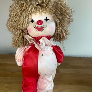 Könnte beinhalten: Eine Vintage-Clown-Puppe mit einem Plüschgesicht, lockigem braunem Haar und einer roten Nase. Das Outfit der Puppe ist geteilt, eine Seite rot und die andere rosa mit bunten Sternen. Der Clown hat einen Rüschenkragen und ist auf einem weißen Sockel montiert.