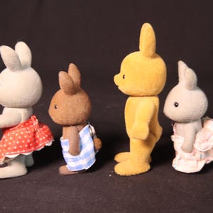 Może przedstawiać: Cztery figurki kr&oacute;lik&oacute;w zabawek w r&oacute;żnych kolorach, każda w unikalnym stroju. Kr&oacute;liki są jasnoszare, brązowe i ż&oacute;łte. Jeden ma na sobie czerwoną sp&oacute;dnicę w białe grochy, drugi niebiesko-biały ręcznik w paski.