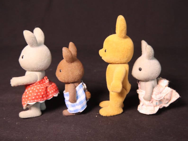 Może przedstawiać: Cztery figurki kr&oacute;lik&oacute;w zabawek w r&oacute;żnych kolorach, każda w unikalnym stroju. Kr&oacute;liki są jasnoszare, brązowe i ż&oacute;łte. Jeden ma na sobie czerwoną sp&oacute;dnicę w białe grochy, drugi niebiesko-biały ręcznik w paski.