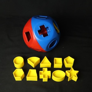 Puede incluir: Un juguete clasificador de formas de plástico multicolor que presenta un gran contenedor esférico rojo y azul con varios recortes geométricos. Diez formas de plástico amarillo distintas están dispuestas en dos filas debajo de la esfera, incluyendo un círculo, triángulo, cuadrado, pentágono, hexágono, trapecio, cruz y estrella.