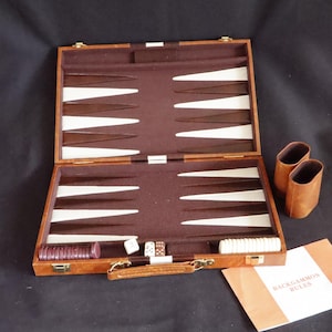 Puede incluir: Juego de backgammon abierto con estuche de cuero marrón, con puntas de juego blancas y marrones. Incluye dados, fichas y un libro de reglas. También se ven dos cubiletes de dados de cuero marrón.