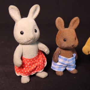 Może przedstawiać: Dwie figurki zabawek kr&oacute;lika. Jedna jest szara i ma na sobie czerwoną sp&oacute;dnicę w kropki. Druga jest brązowa i ma na sobie niebiesko-białe szorty w kratę. Oboje mają czarne oczy i wyprostowane uszy. Tło jest czarne.