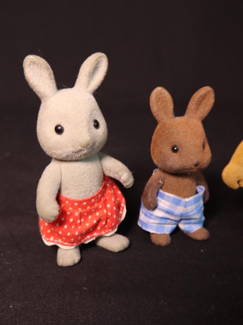 Może przedstawiać: Dwie figurki zabawek kr&oacute;lika. Jedna jest szara i ma na sobie czerwoną sp&oacute;dnicę w kropki. Druga jest brązowa i ma na sobie niebiesko-białe szorty w kratę. Oboje mają czarne oczy i wyprostowane uszy. Tło jest czarne.