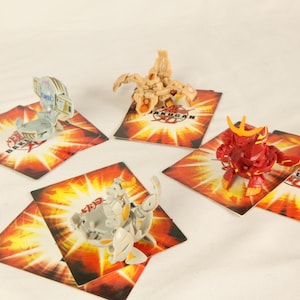 Op de afbeelding: Vier Bakugan-figuren in verschillende kleuren, waaronder rood, beige en grijs, worden getoond op bijpassende speelkaarten. De kaarten hebben een vurig oranje en geel ontwerp met het Bakugan-logo. Het speelgoed staat tegen een witte achtergrond.